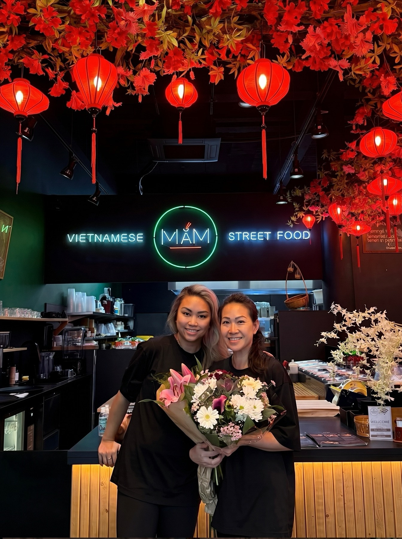 Hiền en Phi-Yến, de twee zussen achter MẮM, met een boeket onder de MẮM-neon